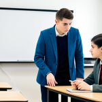A professional coding educator, fully clothed in a smart casual blazer and trousers, gently guiding a young adult student who is also fully clothed in a modest, professional sweater and jeans. They are seated side-by-side at a modern desk in a well-lit, clean tech classroom. The educator is leaning slightly towards the student, pointing at a line of code on the screen with a natural pose, while the student looks attentively at the screen. High-quality professional photography, perfect anatomy, correct proportions, well-formed hands, proper finger count, natural body proportions, sharp focus, vibrant colors, clear composition. Safe for work, appropriate content, fully clothed, professional, modest clothing.